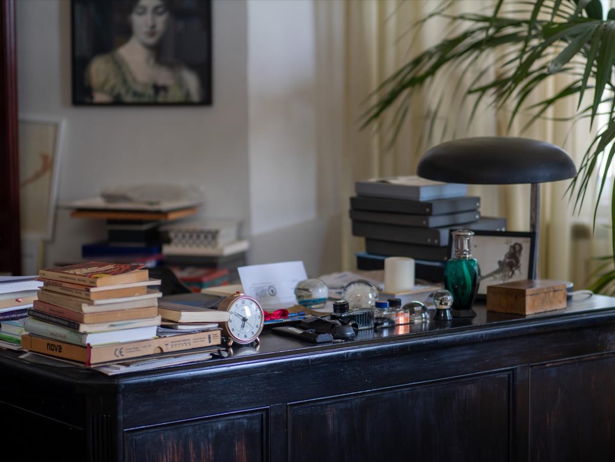 Der schwarze Schreibtisch mit Büchern, Wecker, grünem Flakon und Lampe, dahinter Vorhänge und eine Palme