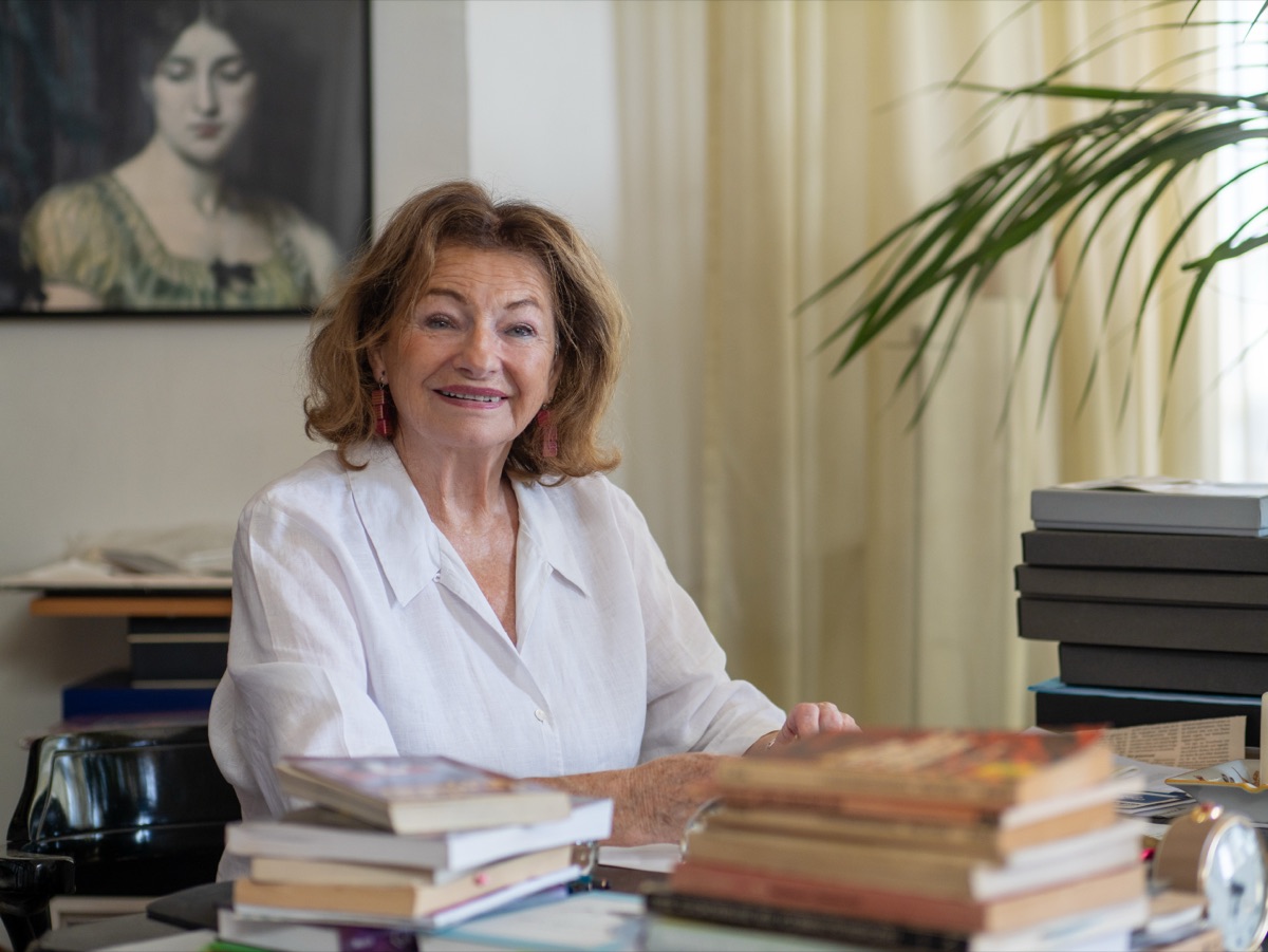 Ingrid Kösten lächelt an ihrem Schreibtisch, zwischen Bücherstapeln, dahinter ein Frauenportrait und eine Palme